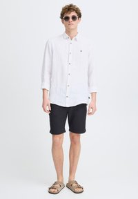 Camisa blanca de lino con botones de madera, combinada con pantalones cortos negros y sandalias marrones. Un hombre se sostiene de pie con confianza, las gafas de sol añaden un detalle.