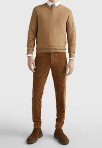 Bruin gewatteerd sweater met ronde hals, gecombineerd met bruine ribfluweel broek met voorzakken en enkellange bruine laarzen.