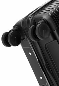 Valise rigide noire équipée de deux roues noires, avec une surface texturée à rainures verticales et une fermeture à glissière sur le bord.