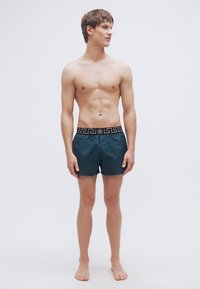 Ανδρικό μαγιό βερμούδα, σε σταθερό σκούρο τιρκουάζ χρώμα με υφασμένο μαύρο λάστιχο στη μέση που φέρει γεωμετρικό σχέδιο. Το ύφασμα φαίνεται ομαλό και ελαφρύ.