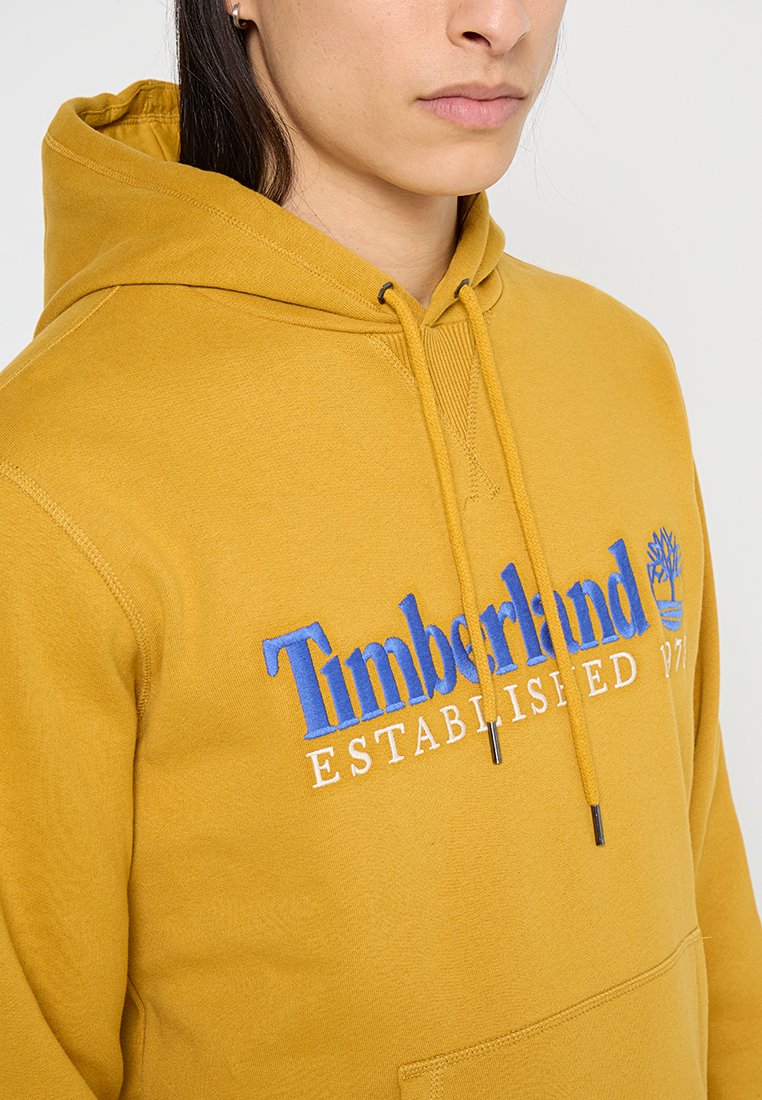 Felpa gialla in cotone con tasca a marsupio frontale, cappuccio regolabile con cordino e logo ricamato "Timberland Established 1973" in blu e bianco.