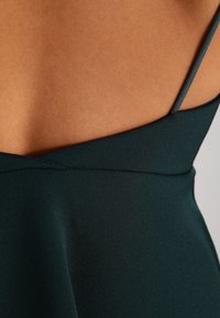 Tumma vihreä mekko, jossa on sileä, mattapintainen tekstuuri. Mekossa on ohuet olkaimet ja V-muotoinen selkädesign. Siisti ompelutaito on näkyvissä.