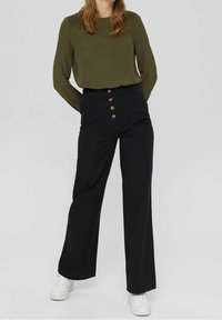 Pantaloni a gamba larga neri a vita alta con apertura frontale a bottoni, abbinati a una camicetta verde oliva a maniche lunghe, con pieghe sulle spalle e texture liscia.