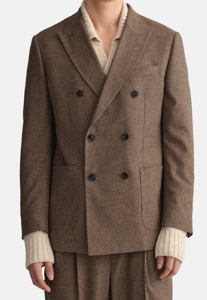 Blazer doppiopetto a quadri marrone con due tasche frontali e bottoni neri, abbinato a un maglione in knit a coste color crema sotto.