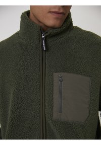 Giacca in pile verde con zip e colletto alto, caratterizzata da un tessuto testurizzato e una tasca sul petto con zip in un materiale più liscio.