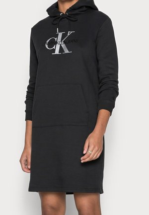 Czarna sukienka bluza z kapturem Calvin Klein z przednią kieszenią i logo na piersi, noszona przez osobę z pierścionkiem na prawej ręce.