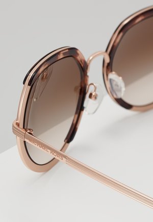 Primer plano de gafas de sol de color oro rosa y carey con lentes degradadas marrones y el logo de Michael Kors en la patilla.