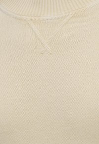Gros plan sur un tissu tricoté couleur crème avec un col côtelé et un détail décoratif en V cousu sous l'encolure.