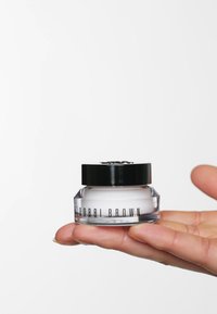 Klares Glasgefäß mit einem schwarzen Deckel, das eine weiße Creme enthält. Etikett lautet "BOBBI BROWN". In einer Hand vor einem weißen Hintergrund gehalten.