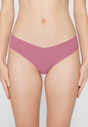 Weiblicher Oberkörper mit nahtloser Bikini-Unterwäsche in Mauve, Hände locker an den Seiten, schlichter heller Hintergrund.