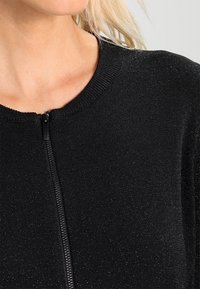 Maglione nero con zip e scollo rotondo, caratterizzato da una finitura testurizzata scintillante e polsini a costine, adatto a look stratificati.