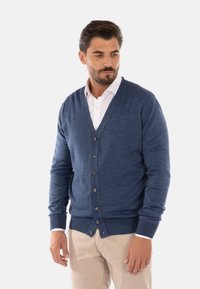 Cardigan azzurro a scollo a V realizzato in tessuto knit leggero. Presenta chiusura con bottoni, polsini e orlo a costine. Indossato sopra una camicia bianca.
