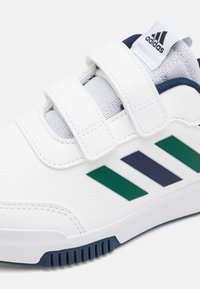 Białe sportowe buty z zapięciami na rzep, z zielonymi i granatowymi paskami, teksturowanym materiałem i gumową podeszwą. Zaprojektowane z myślą o komforcie i wsparciu.