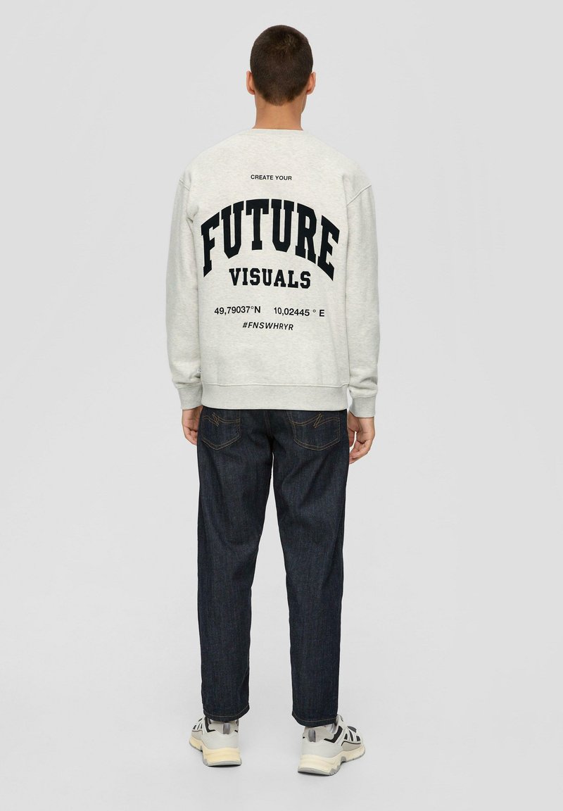 Hellgrauer Pullover mit schwarzem Text auf dem Rücken, der "Erschaffe deine zukünftigen Visuals" liest, kombiniert mit dunklen Jeans und Turnschuhen.