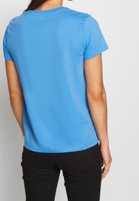 Hellblaues Baumwoll-T-Shirt mit kurzen Ärmeln, Rundhalsausschnitt und schlichter Textur, von hinten vor einem neutralen Hintergrund gezeigt. Schwarze Hose sichtbar.