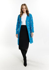 Blå, texturerad cardigan med paljetter, över en vit topp och svart kjol, kombinerad med höga svarta stövlar. Enkel design med öppen front.