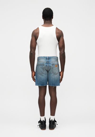 Džínové kraťasy s prechodom farby od svetlomodrej po tmavomodrú, s ošúchanými okrajmi a zadnými vreckami, nosené s bielym tielkom, čiernymi topánkami a bielymi ponožkami.