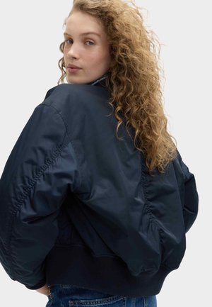 Junge Frau mit langen lockigen Haaren, die eine lockere dunkelblaue Bomberjacke und Jeans trägt und sich leicht zurückdreht vor schlichtem Hintergrund.