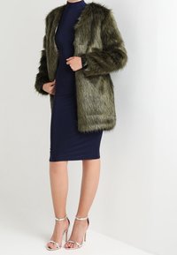 Olivgrön fuskpälsrock med texturerad yta, öppen fram, och långa ärmar, bärs över en åtsittande marinblå klänning och silvriga klacksandaler.