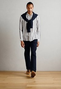 Helle gestreifte Hemd, dunkler Pullover um die Schultern gebunden, dunkle Hose und helle Schuhe. Glatte Texturen, lässiges Design.