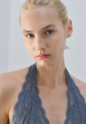 Mujer rubia con ojos azules que lleva un top halter de encaje azul, mirando directamente a la cámara con una expresión neutral sobre un fondo liso.