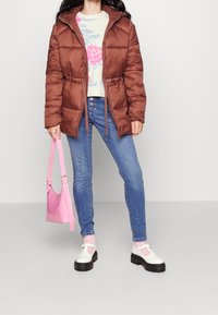 Braune Steppjacke mit Kordelzügen, kombiniert mit einem hellen Blumensweater. Blaue Jeans und rosa Tasche. Weiß-rosa Schuhe mit dicken schwarzen Sohlen.