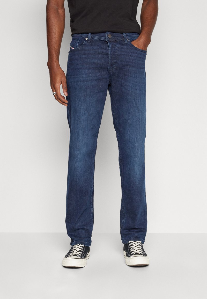 Jeans din denim de culoare bleumarin, cu croială dreaptă, având buzunare frontale, bucle pentru curea și detalii de cusătură subtile. Purtați cu adidași negri.