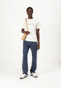 Hvit bomull t-skjorte med trykt logo, blå denimbukser, beige crossover-veske og grå joggesko med svarte striper. Enkel, uformell antrekk.