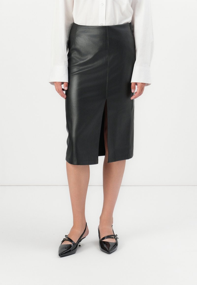 Femme portant une jupe midi en cuir noir avec une fente devant, une chemise blanche à manches longues et des talons slingback noirs à bout pointu.