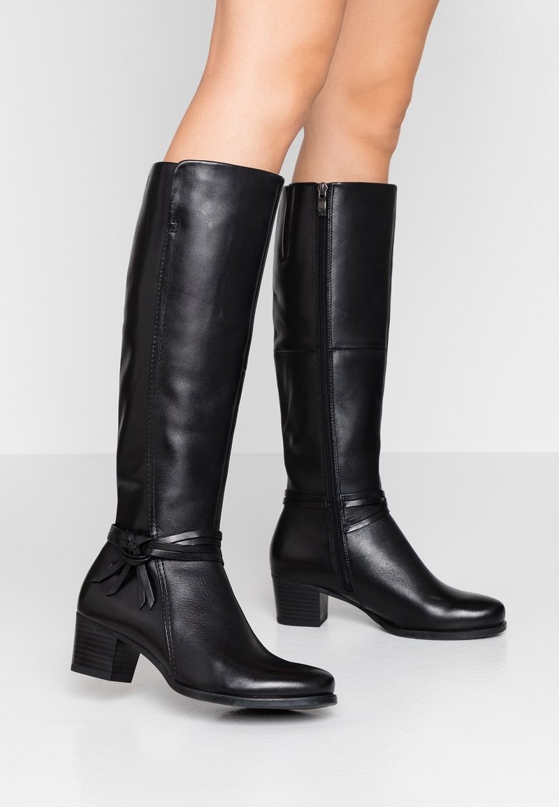 Botas de cuero negro hasta la rodilla con tacón grueso, cremallera lateral y detalles decorativos de lazos en el tobillo. Textura suave y diseño elegante.