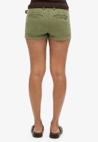 Shorts en coton verts avec une coupe ajustée, dotés de deux poches arrière et d'un ourlet replié, associés à des sandales noires.