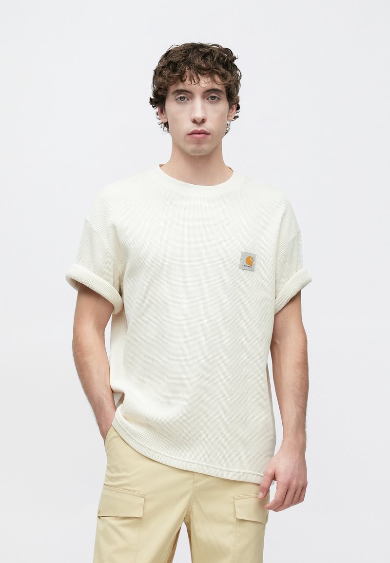 Off-white, ribbed short-sleeve t-shirt with rolled cuffs and a square patch logo on the chest; paired with light beige cargo pants.