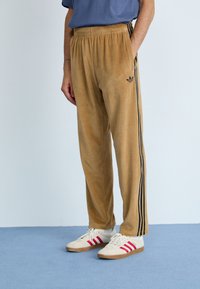 Calças de veludo castanho com riscas laterais pretas e um logótipo bordado na perna esquerda, combinadas com ténis brancos com detalhes em vermelho.