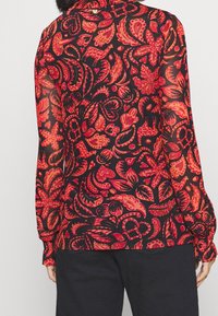 Blusa nera trasparente con motivo floreale rosso e arancione. Maniche lunghe con polsini arricciati, dettaglio colletto e delicati dettagli metallici.