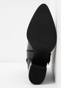 Chaussure habillée en cuir noir reposant sur le talon, montrant la semelle pointue et le détail de la bride sur fond blanc.