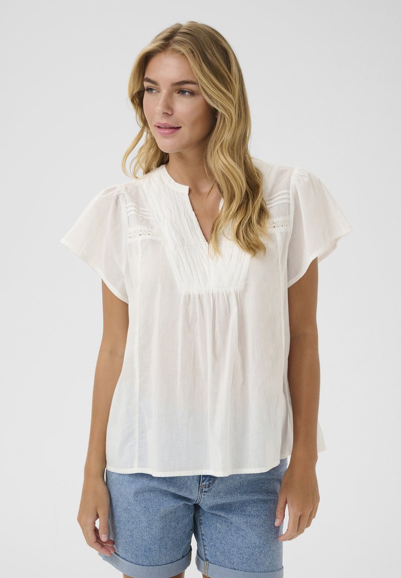 Blouse blanche légère avec des manches courtes flutter, un col rond et des détails plissés. Le tissu semble doux avec une texture subtile.