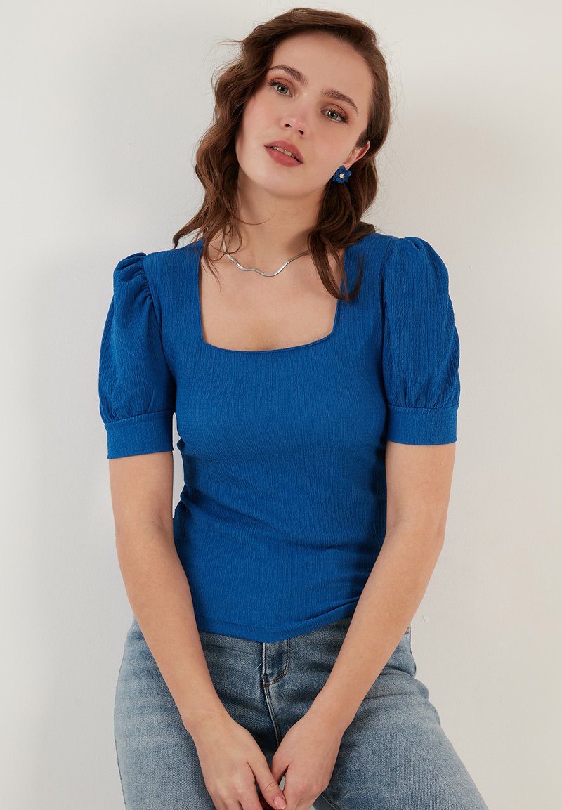Maglietta aderente blu royal con scollatura quadrata, maniche a sbuffo corte, tessuto testurizzato, indossata con jeans blu chiaro. Collana semplice in argento come accentuazione.