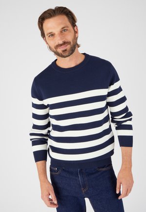 Maglione a righe blu navy e bianche con scollo rotondo, polsini e orlo a costine. Indossato con jeans in denim scuro. Texture a maglia liscia.
