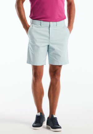 Mann trägt hellblaue, maßgeschneiderte Shorts, dunkelblaue Turnschuhe und ein rosa Hemd, steht mit den Händen in den Taschen vor weißem Hintergrund.