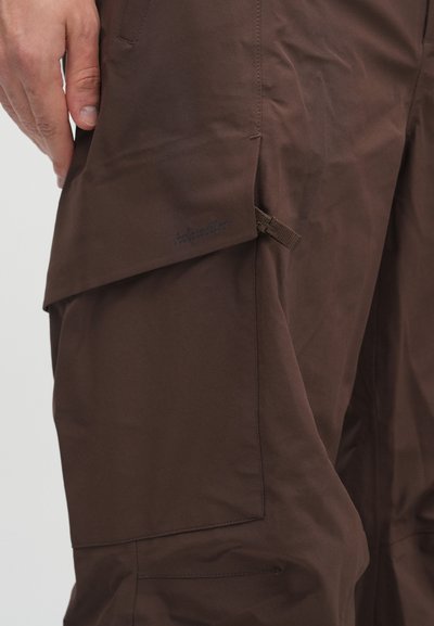 Rjave cargo hlače, izdelane iz gladkega materiala, z velikimi stranskimi žepi in diskretno logo. Oblikovanje poudarja uporabnost in udobje.