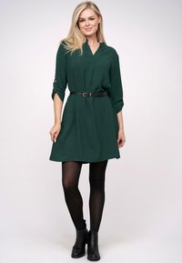 Grünes Kleid mit V-Ausschnitt, dreiviertelÄrmeln und einem schwarzen Gürtel. Das Kleid hat eine sanfte Textur und eine ausgestellte Form, kombiniert mit schwarzen Stiefeletten.