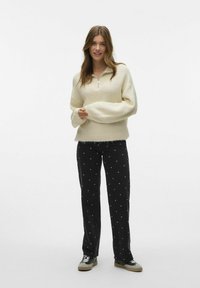 Kremfarget, zip-up genser med vide ermer, kombinert med svarte bukser med hvite prikkemønstre. Fottøyet inkluderer svarte og grå sneakers.