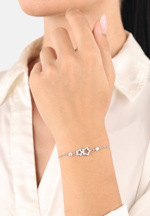 Delicata bracciale d'argento con due pendenti a forma di stella e piccole pietre tonde indossato su un polso, abbinato a una blusa bianca in raso.