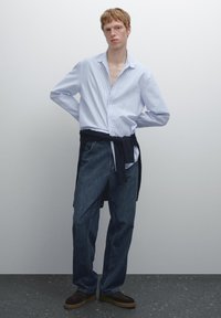 Langarm gestreept shirt met een open kraag, gecombineerd met donkerblauwe jeans. Marineblauwe trui om de taille geknoopt en bruine schoenen.