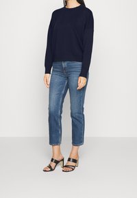 Una donna con un maglione blu marino, jeans blu a vita alta e sandali neri con tacco, in piedi davanti a uno sfondo chiaro e uniforme.