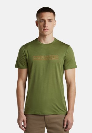 Junger Mann mit kurzen Haaren, der ein olivgrünes T-Shirt mit kurzen Ärmeln und dem Text "ICEBREAKER" sowie Khakihosen trägt, steht vor einem weißen Hintergrund.