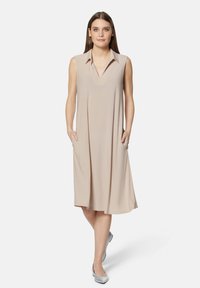 Ärmelloses beige Kleid mit spitzem Kragen, V-Ausschnitt und seitlichen Taschen; aus glattem Stoff, der sanft bis zum Knie fließt.