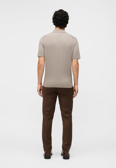 Homme aux cheveux bouclés portant un polo beige à manches courtes, un pantalon marron et des chaussures marron, debout de dos sur un fond uni.