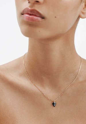 Primer plano del cuello y la parte inferior del rostro de una persona que lleva un delicado collar de cadena dorada con un pequeño colgante en forma de punta de flecha negra.