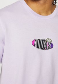 Detailní záběr osoby v světle fialové košili s barevným logem Adidas na hrudi.